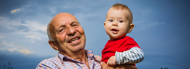 Grandfather holding grandson outdoors, against a sunset sky, smiling