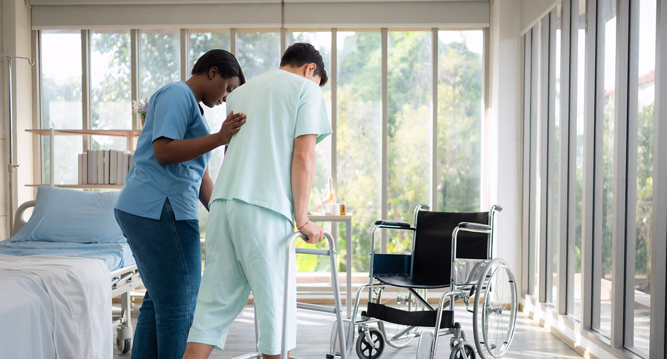 Young female nurse helping young male patient to walk on walker at patient room